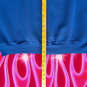 May include: A close-up of the bottom hem of a blue sweatshirt. A measuring tape is visible on the right side of the image.