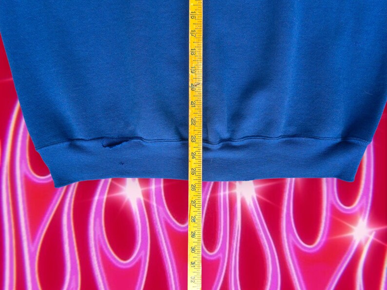 May include: A close-up of the bottom hem of a blue sweatshirt. A measuring tape is visible on the right side of the image.