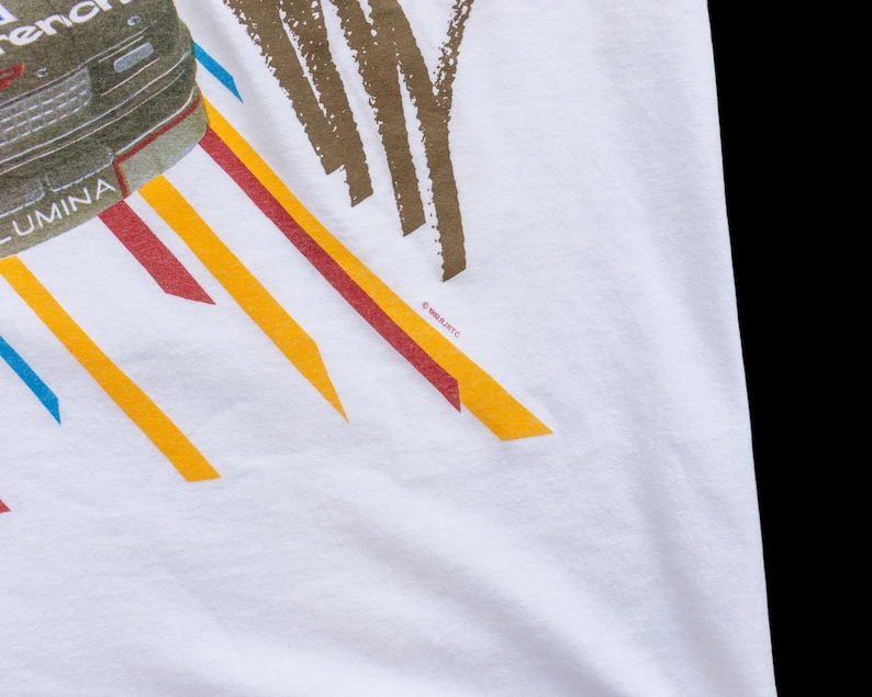 May include: White t-shirt with a graphic of a black car with the word "LUMINA" on the side. The car is surrounded by colorful stripes and the text "&copy;1991 GM/ITC" is visible in the bottom right corner.