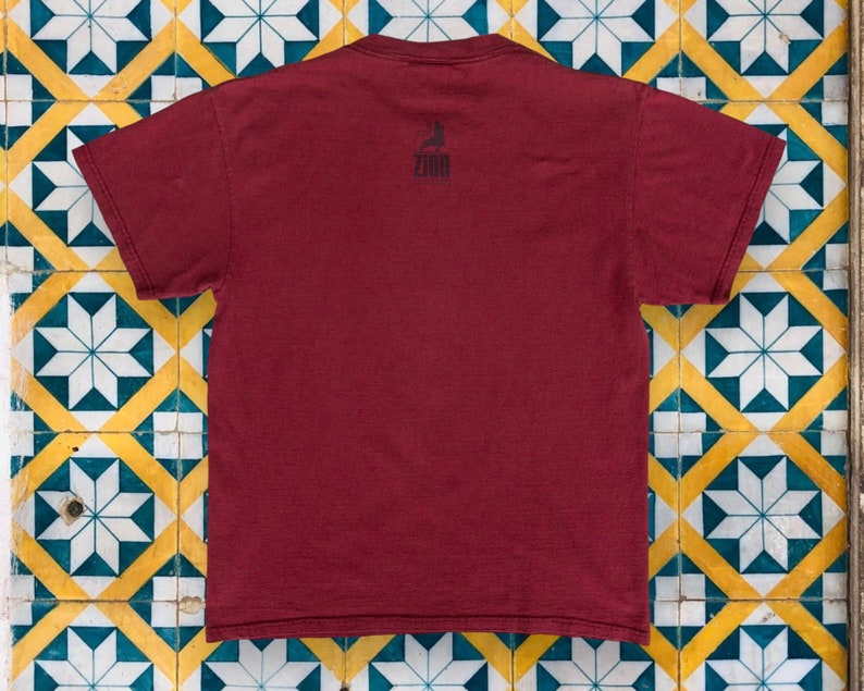 May include: A maroon t-shirt with a black graphic of a person surfing on a wave. The graphic includes the word "ZOO" in black text.