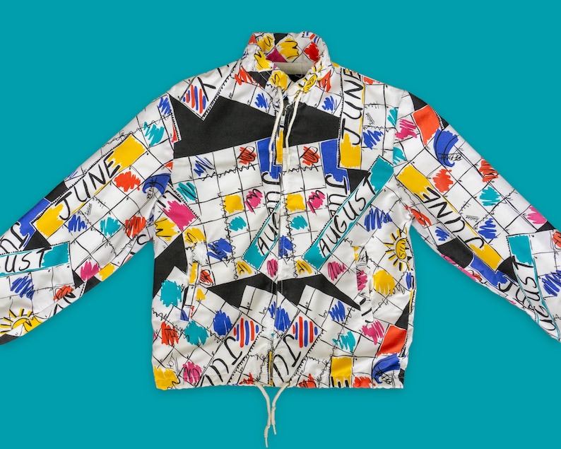 Op de afbeelding: Een wit, zwart, geel, blauw en roze gepatroneerd jack met een ritssluiting. Het patroon bevat de maanden juni en augustus.