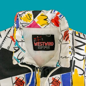 Op de afbeelding: Een wit en zwart ritsjack met een kleurrijke abstracte print. Het jack heeft een witte kraag en een zwarte rits. Het label op het jack luidt "Westwind Sportswear Made in USA."