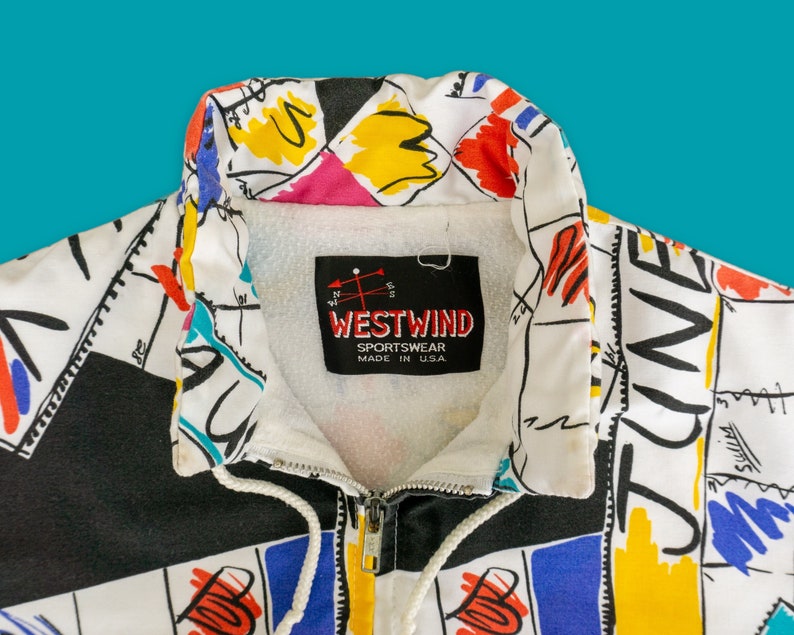 Op de afbeelding: Een wit en zwart ritsjack met een kleurrijke abstracte print. Het jack heeft een witte kraag en een zwarte rits. Het label op het jack luidt "Westwind Sportswear Made in USA."
