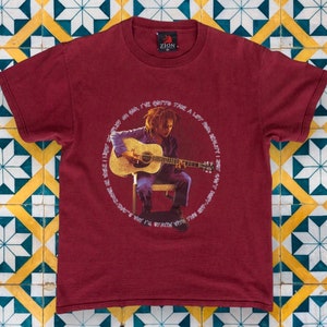 May include: A maroon t-shirt with a graphic print of a man playing a guitar. The text around the image reads "I've gotta take a lot of red, really I don't want to be dancing, why I'm staying with the rest of you".
