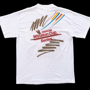 May include: White t-shirt with a colorful graphic design on the back. The graphic features a stylized eagle, the text "NASCAR Winston Cup Series", and abstract lines in brown, red, blue, and yellow.