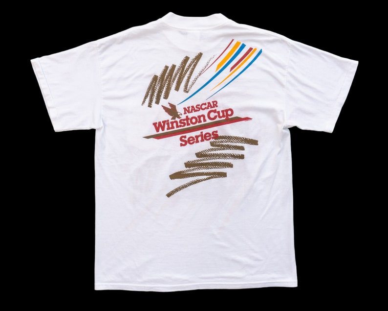 May include: White t-shirt with a colorful graphic design on the back. The graphic features a stylized eagle, the text "NASCAR Winston Cup Series", and abstract lines in brown, red, blue, and yellow.