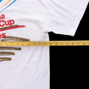 May include: White t-shirt with a red, white, and blue NASCAR Winston Cup Series logo. The logo is printed on the front of the shirt. The shirt is laid flat on a surface with a measuring tape.