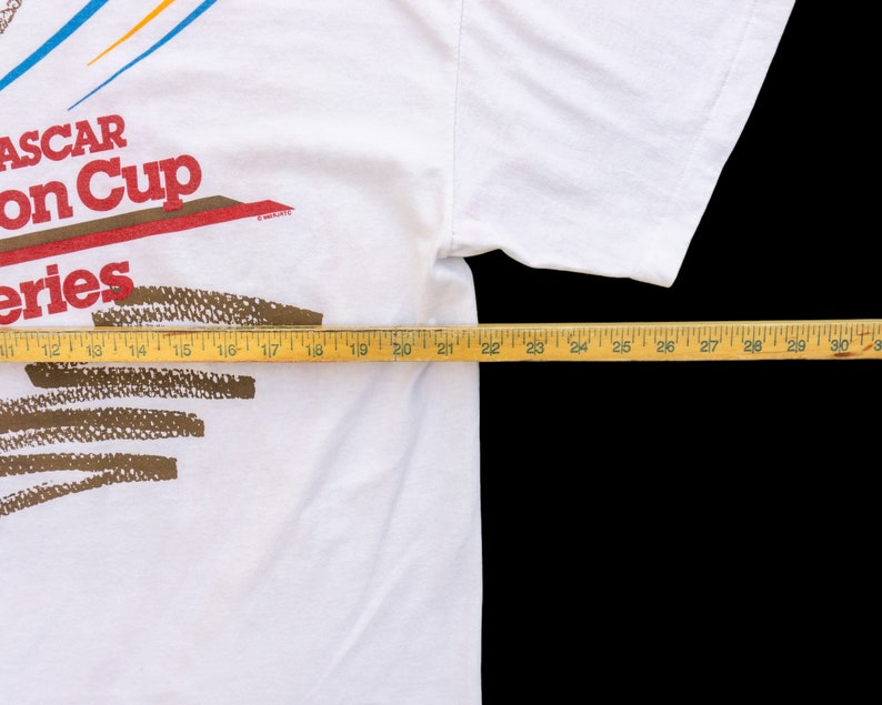 May include: White t-shirt with a red, white, and blue NASCAR Winston Cup Series logo. The logo is printed on the front of the shirt. The shirt is laid flat on a surface with a measuring tape.