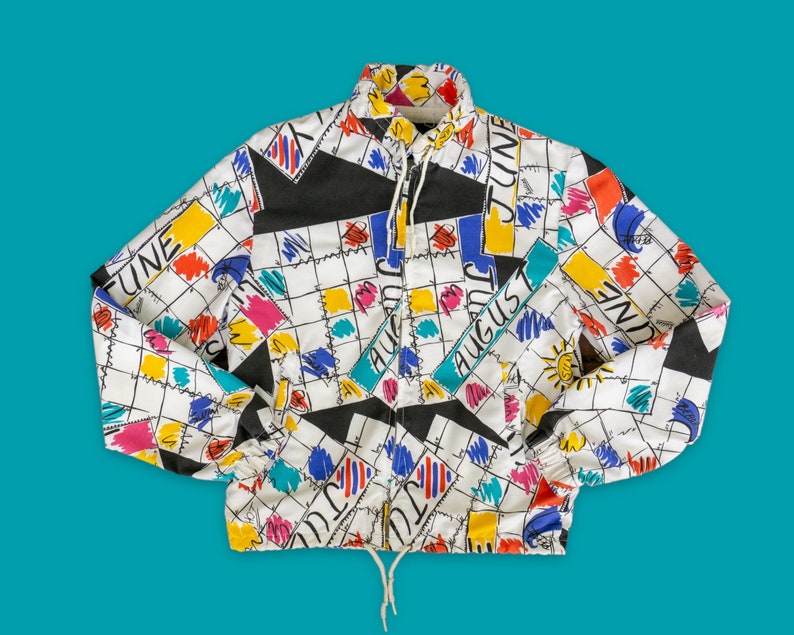 Op de afbeelding: Een wit, zwart, blauw, geel en rood gepatroneerd jack met een ritssluiting. Het jack heeft een kalenderthema met de woorden "juni", "juli" en "augustus" erop gedrukt.