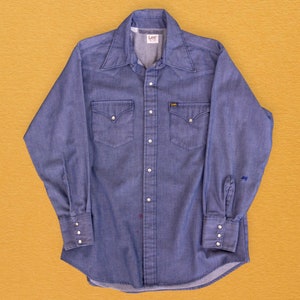 May include: A blue denim long-sleeve shirt with a button-down collar, two chest pockets, and the word "Lee" on the left side of the collar.