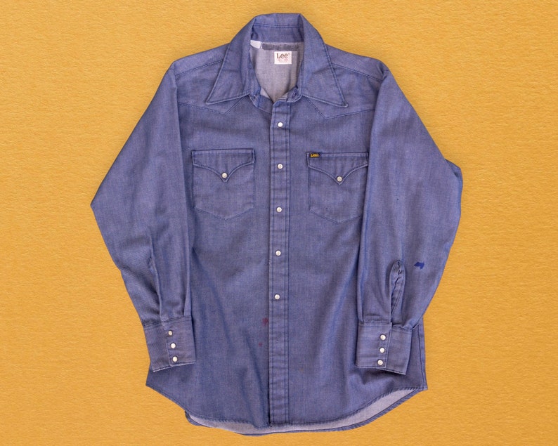 May include: A blue denim long-sleeve shirt with a button-down collar, two chest pockets, and the word "Lee" on the left side of the collar.