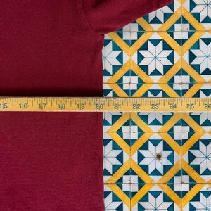 May include: A close-up of a maroon t-shirt with a wooden ruler measuring 20 inches in length. The ruler is placed on a tiled surface with a geometric pattern of white, yellow, and blue.