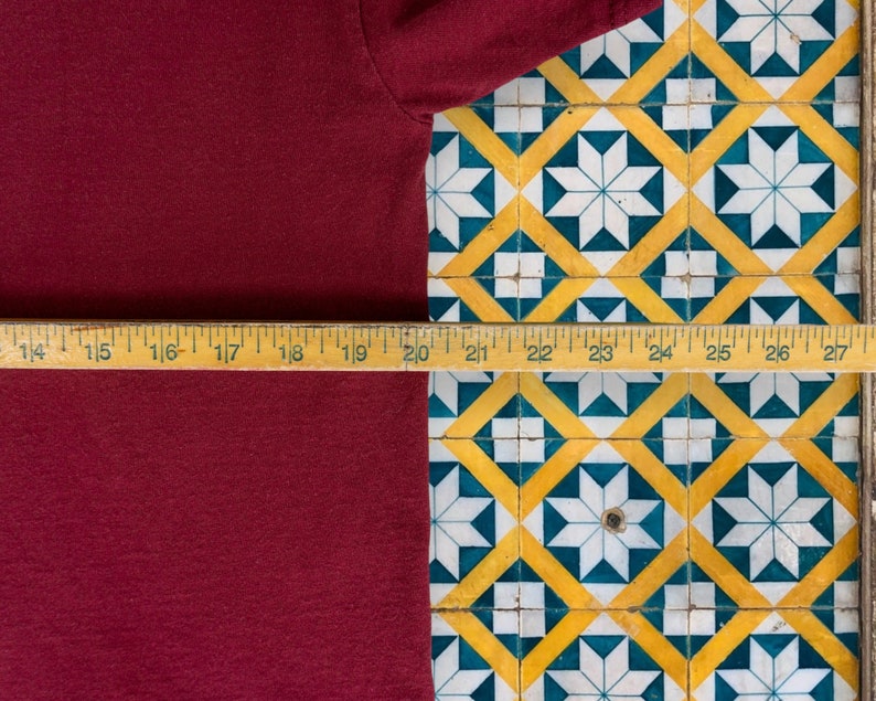 May include: A close-up of a maroon t-shirt with a wooden ruler measuring 20 inches in length. The ruler is placed on a tiled surface with a geometric pattern of white, yellow, and blue.