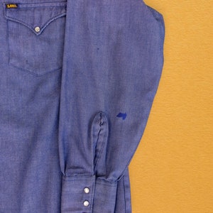 May include: A blue denim long-sleeve shirt with a single chest pocket and a Lee logo on the left side. The shirt has a button-down collar and button cuffs.