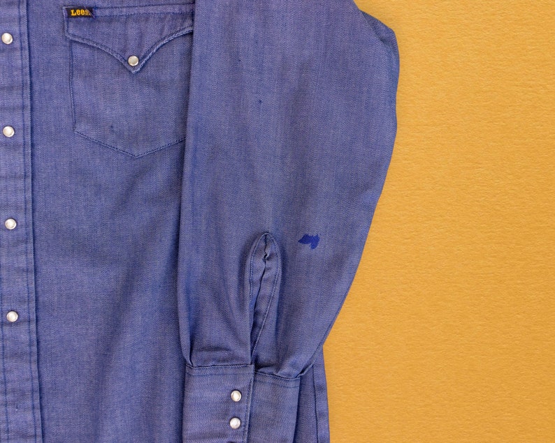 May include: A blue denim long-sleeve shirt with a single chest pocket and a Lee logo on the left side. The shirt has a button-down collar and button cuffs.