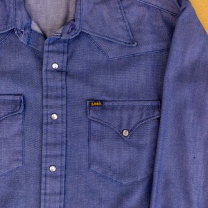 May include: A blue denim long-sleeve shirt with a pointed collar, button closure, and two chest pockets. The shirt has a Lee brand label on the left chest pocket.