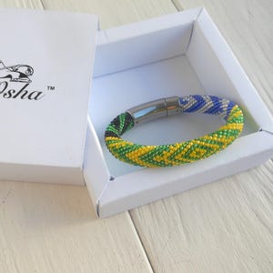 May include: A colourful beaded bracelet with yellow, green, blue, and white sections sits in a white box. The bracelet has a magnetic clasp. The box features the text "ifa-Osha" and a lion logo.