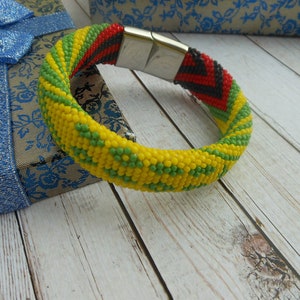 May include: A beaded bracelet in yellow, green, red, and black. The bracelet has a magnetic clasp and is displayed on a wooden surface next to a gift box with a blue bow. The bracelet is a vibrant accessory.
