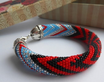 Può includere: Un braccialetto di perline con un motivo geometrico in rosso, nero, argento e blu. Il braccialetto ha una chiusura argentata.