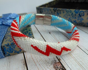 Può includere: Un braccialetto di perline con chiusura magnetica. Il braccialetto presenta un motivo di perline bianche, rosse e azzurre. Il design include un motivo a zig-zag rosso. Il braccialetto è su una superficie di legno.