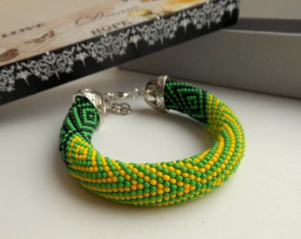 May include: A green, yellow, and black beaded bracelet with a silver clasp. The bracelet is made of small, round beads that are woven together in a geometric pattern.