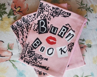 May include: Pink canvas zippered pouch with a black and white graphic print and the text "BURN BOOK" in black letters with a red lip print.