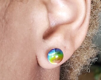 May include: A circular stud earring featuring a smooth, domed surface with a vibrant gradient of colours transitioning from blue at the top to green and yellow at the bottom. The earring has a polished, reflective finish and is worn in the earlobe.