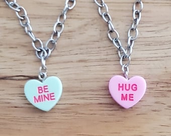 Op de afbeelding: Vier zilveren kettingen met hartvormige bedels. De bedels zijn pastelroze, lichtblauw en wit met de woorden "Hug Me", "Be Mine" en "Be Mine" in roze.