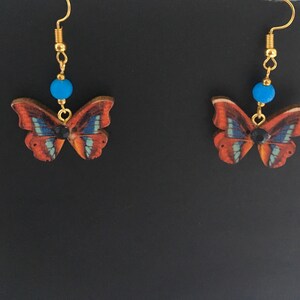 May include: A pair of gold butterfly earrings with blue beads and black accents. The butterflies are made of wood and have a brown, orange, and blue pattern.
