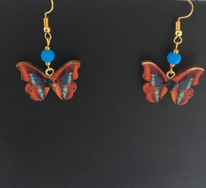 May include: A pair of gold butterfly earrings with blue beads and black accents. The butterflies are made of wood and have a brown, orange, and blue pattern.