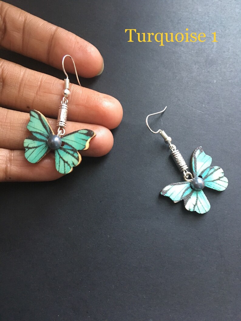 May include: A pair of turquoise butterfly earrings with silver hooks and black pearl accents. The earrings are displayed on a black surface. Turquoise 1.