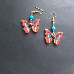 May include: A pair of dangle earrings featuring a butterfly design with red, blue, and white accents. Each earring has a turquoise bead and a black rhinestone.