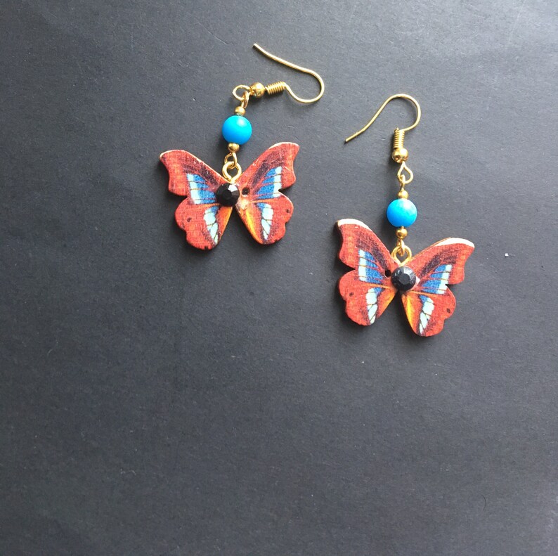 May include: A pair of dangle earrings featuring a butterfly design with red, blue, and white accents. Each earring has a turquoise bead and a black rhinestone.