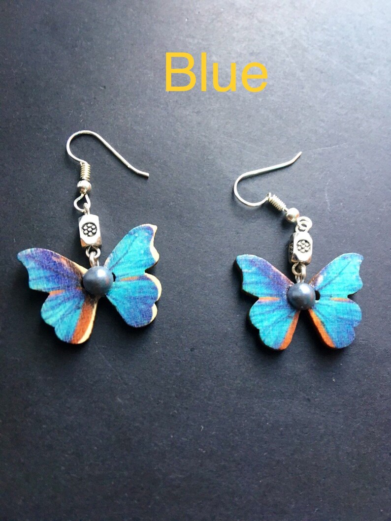 May include: A pair of blue butterfly earrings with silver accents. The butterflies are made of wood and have a blue and orange pattern. The earrings are hanging from silver hooks.