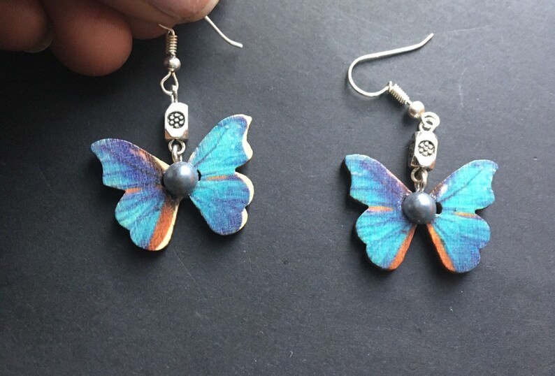 May include: A pair of butterfly earrings with blue and orange wings, silver accents, and a dark gray pearl in the center of each butterfly.