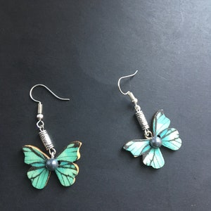 May include: A pair of silver butterfly earrings with a teal and black butterfly design and a dark gray pearl accent.