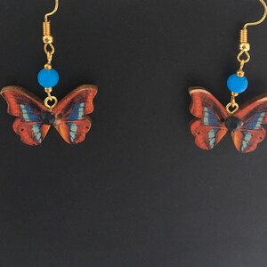 May include: A pair of gold butterfly earrings with blue beads and black accents. The butterflies are made of wood and have a brown, blue, and black pattern.