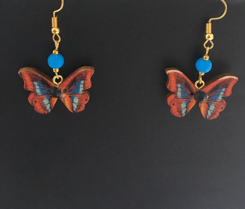 May include: A pair of gold butterfly earrings with blue beads and black accents. The butterflies are made of wood and have a brown, blue, and black pattern.