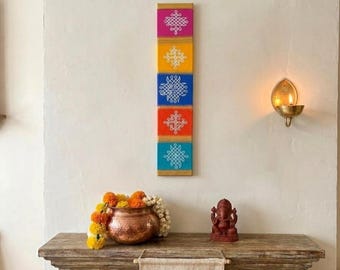 Puede incluir: Una decoración mural vertical con cinco cuadrados de colores: rosa, amarillo, azul, naranja y turquesa. Cada cuadrado tiene un diseño bordado en blanco. La decoración de la pared se muestra encima de una mesa de madera con una olla de cobre, flores y una pequeña estatua.