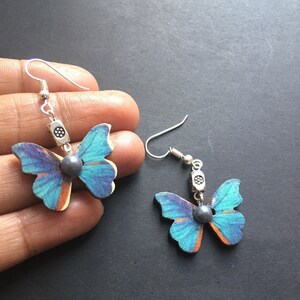 May include: A pair of blue butterfly earrings with silver hooks and a dark gray bead in the center of each butterfly.