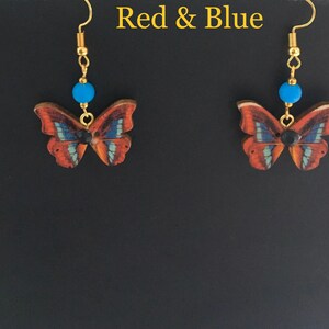 May include: A pair of dangle earrings featuring a butterfly design with red, blue, and black accents. The butterflies are made of wood and have a gold-tone finish. The earrings are attached to gold-tone hooks.