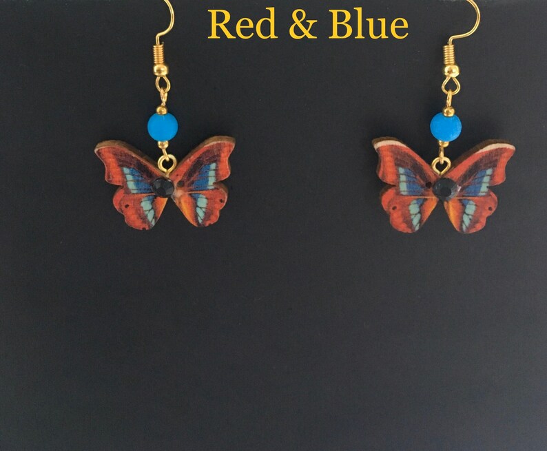 May include: A pair of dangle earrings featuring a butterfly design with red, blue, and black accents. The butterflies are made of wood and have a gold-tone finish. The earrings are attached to gold-tone hooks.