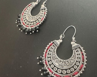 May include: A pair of silver hoop earrings with intricate designs and black beads hanging from the bottom. The earrings have a red thread wrapped around the hoop.