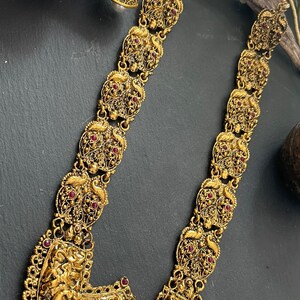Könnte beinhalten: Eine goldfarbene Halskette mit filigranen Details und roten Edelsteinen. Die Halskette hat einen großen Anhänger mit einem detaillierten Design von Figuren. Die Halskette ist mit Perlentropfen verziert.