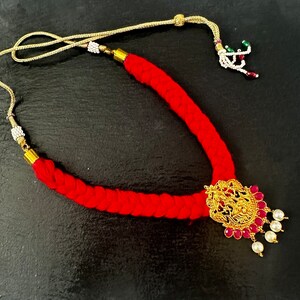 May include: A red braided necklace with a gold pendant featuring a detailed design and red and white stones. The necklace has a gold clasp and a gold tassel with green and white beads.