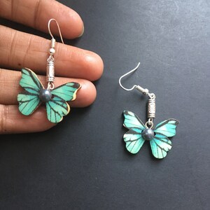 May include: A pair of silver butterfly earrings with a teal and white butterfly design and a black pearl accent.