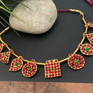 Peut inclure: Un collier doré orné de pendentifs de formes géométriques et de feuilles. Chaque pendentif est orné de petites pierres précieuses rouges et vertes. Le collier est présenté sur un fond sombre, mettant en valeur son design complexe et ses couleurs vives.