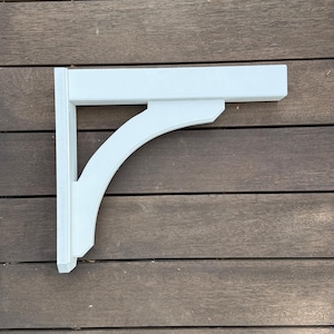 May include: A white wooden corbel with a curved support and a rectangular top. The corbel is designed to provide structural support and decorative detail. The piece is painted white and is set against a wooden background.