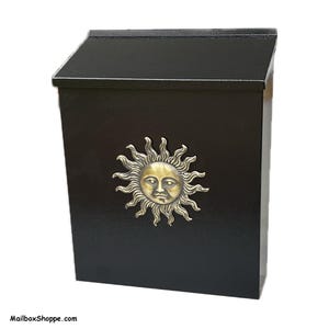 May include: A black wall-mounted mailbox featuring a slanted top lid and a centered, circular brass-toned sun emblem with a human face. The front surface has a textured finish, and the text MailboxShoppe.com is printed in the bottom left corner.