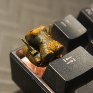 May include: A small, detailed, 3D printed model of a spaceship, painted in a rusty brown and green color scheme, is sitting on a black keyboard.